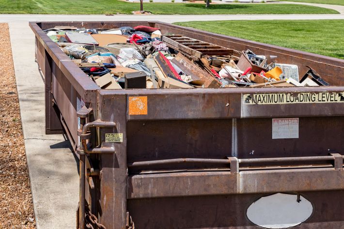 Large brown dumpster filled with construction debris, parked beside a sidewalk and grassy lawn.