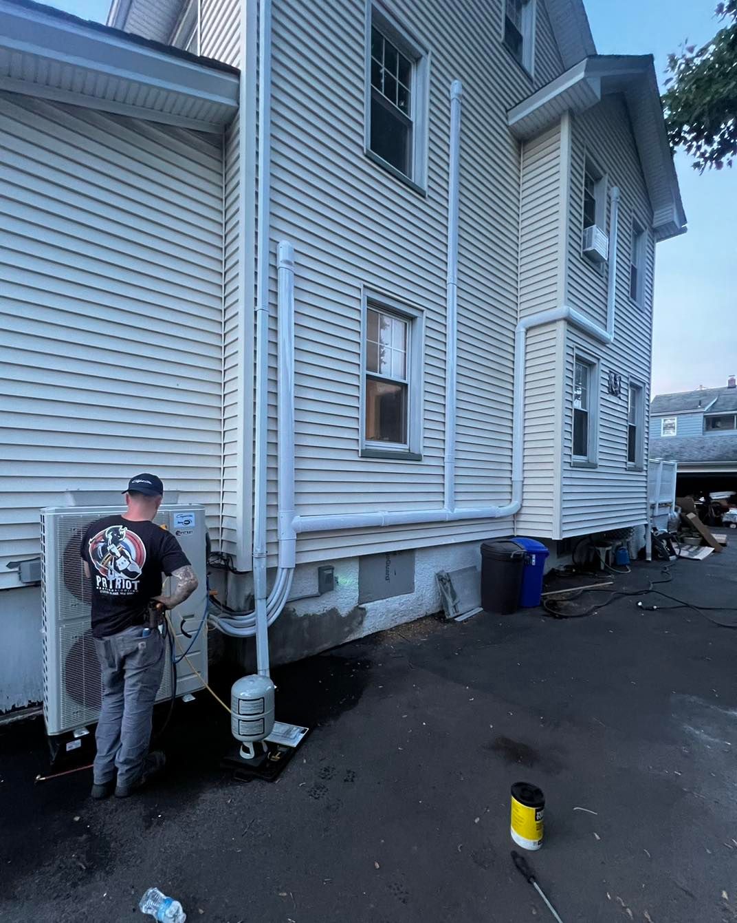 A worker installing an outdoor HVAC unit on a white house exterior with piping.
