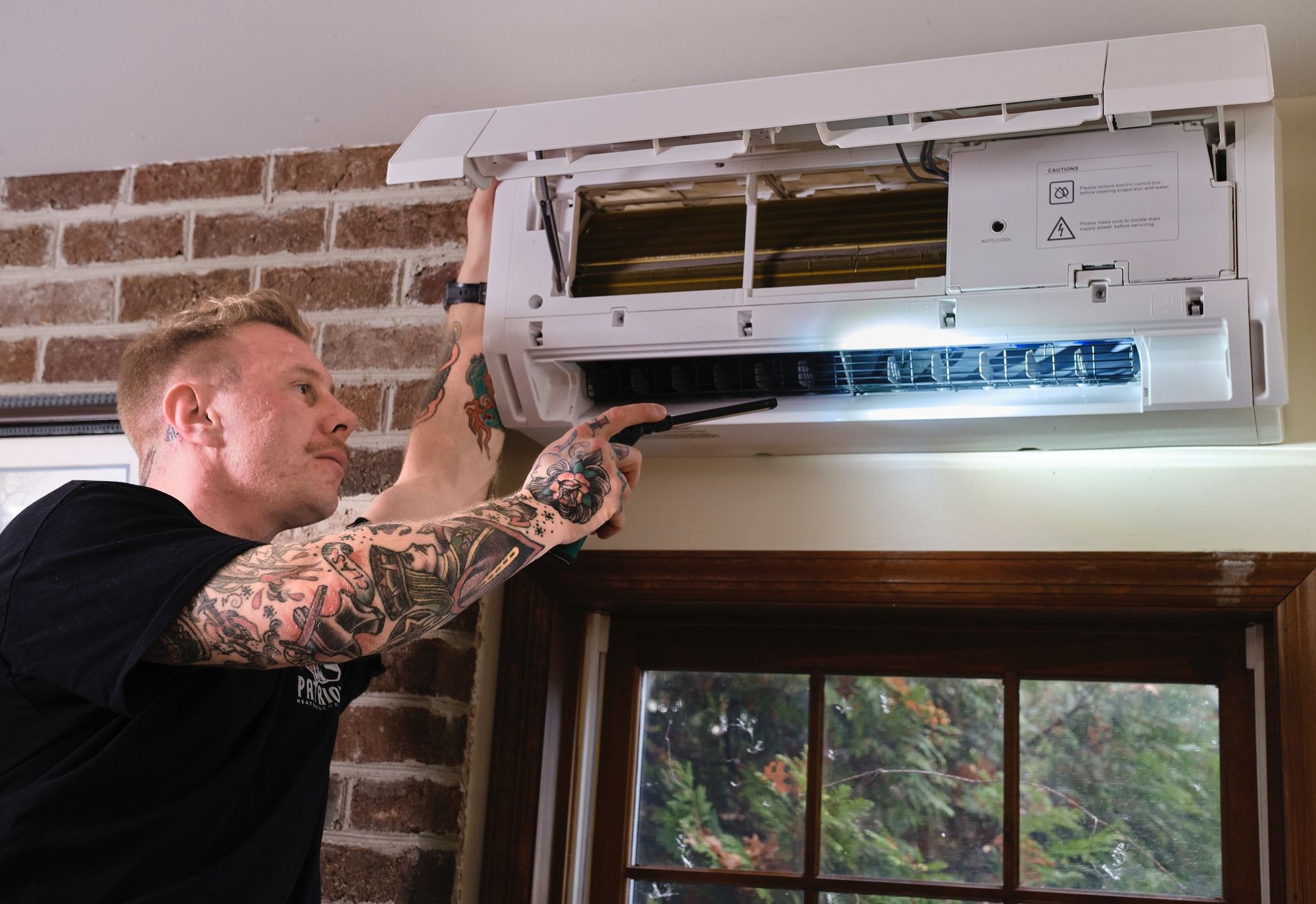 Electrician in yellow hard hat, overalls, black gloves works on electrical panel. Window in background.