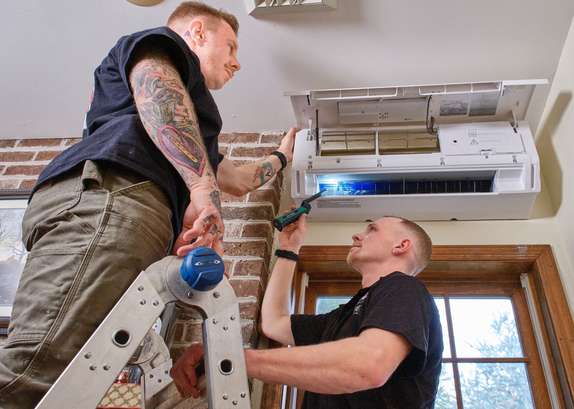 Electrician in yellow hard hat, overalls, black gloves works on electrical panel. Window in background.