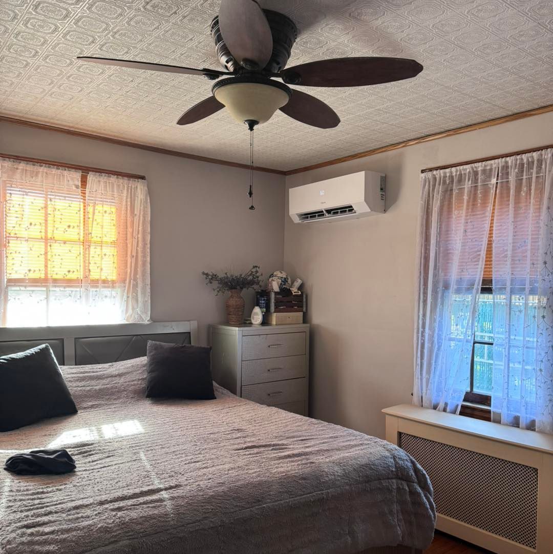 Bedroom with bed, windows, air conditioner, ceiling fan. Light streaming through curtains.