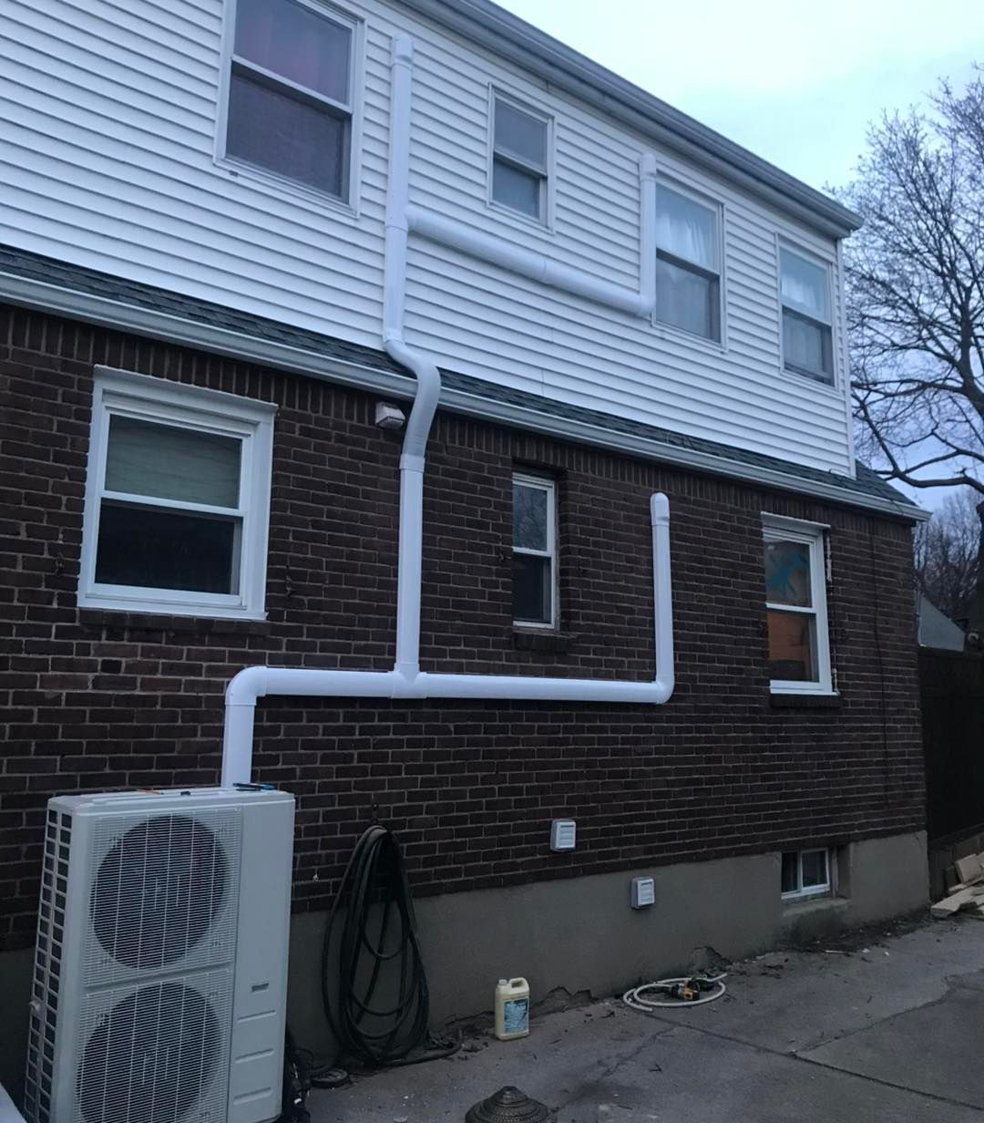 White pipes on a brick building, connected to an outdoor HVAC unit and venting upward. Overcast sky.