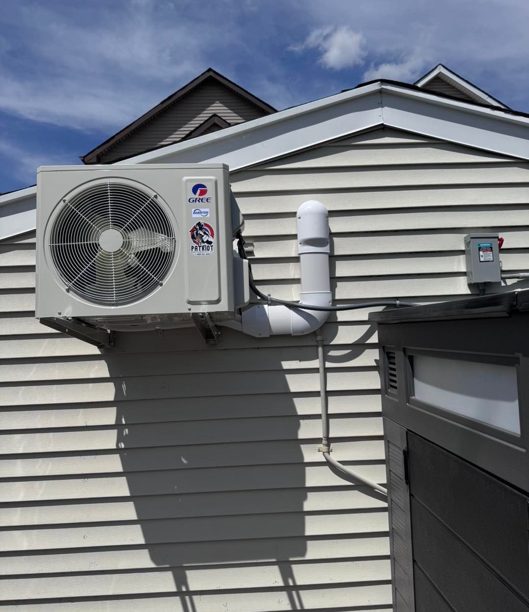 An air conditioning unit mounted on a white clapboard wall with a white pipe and electrical box.