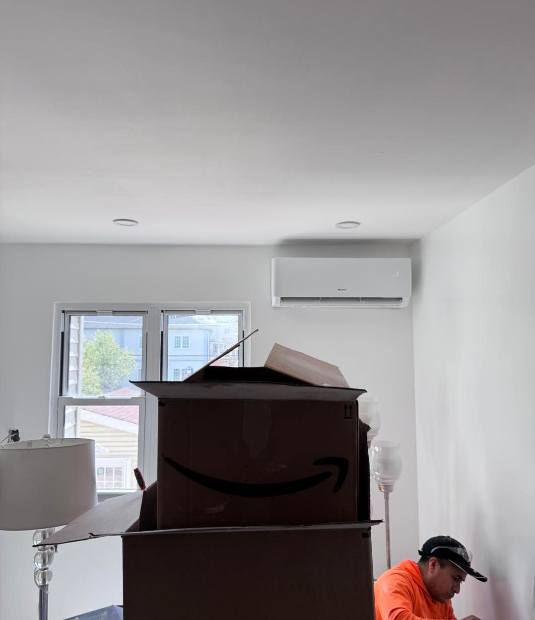 A person unpacking boxes in a room with white walls, window, and air conditioner unit.