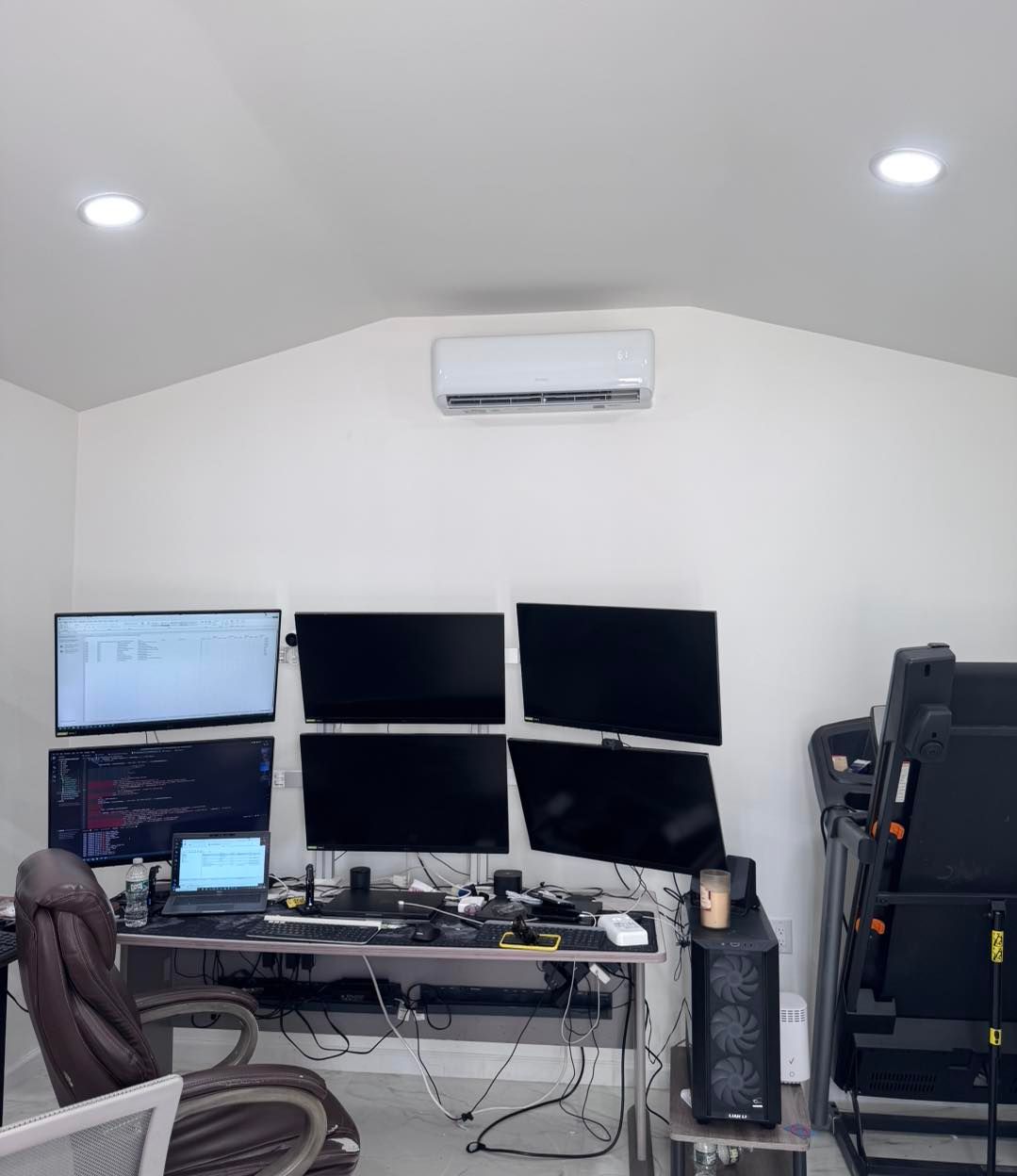 A home office with multiple computer monitors, a desk, a treadmill, and an air conditioner unit.