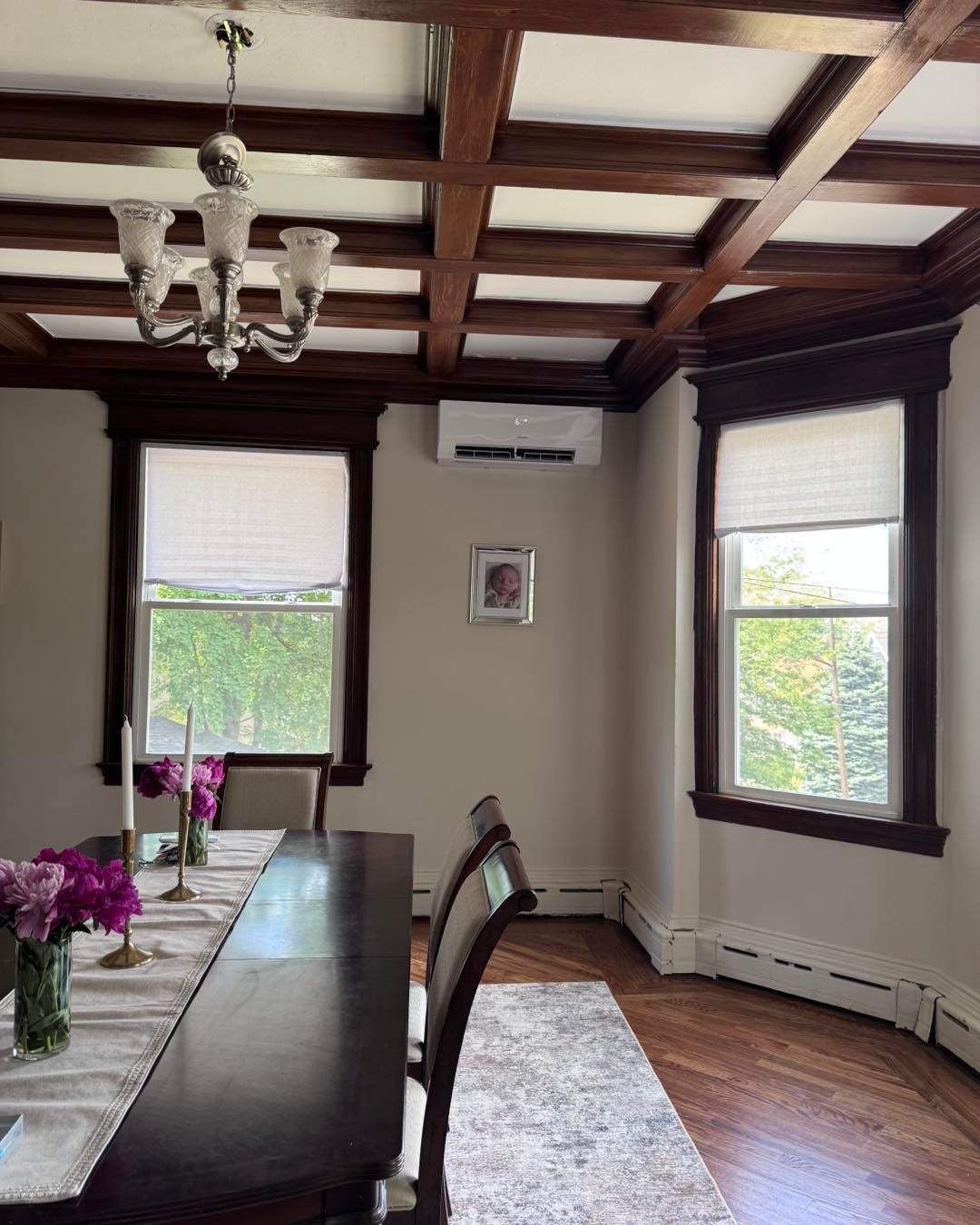 Dining room with dark wood trim, coffered ceiling, windows, chandelier, long table, and an AC unit.