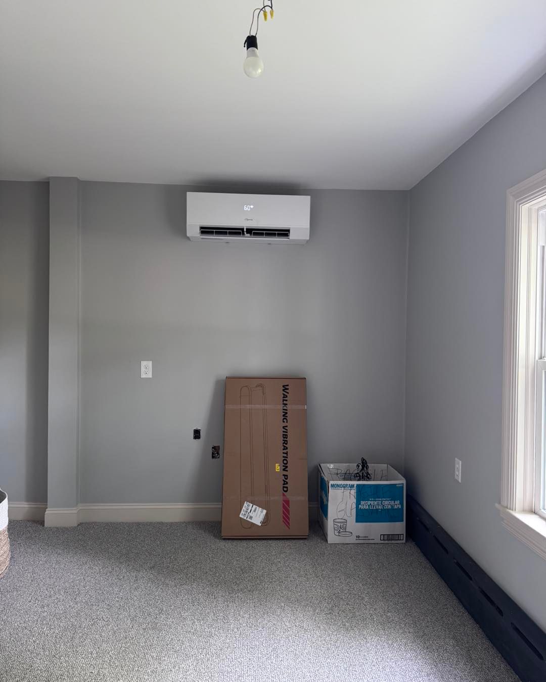 Room with gray walls, mini-split AC, boxes on floor, unfinished flooring, window, and exposed light fixture.