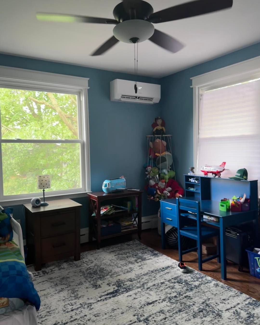 Blue bedroom with windows, a rug, desk, toy shelf, and air conditioner on the wall.