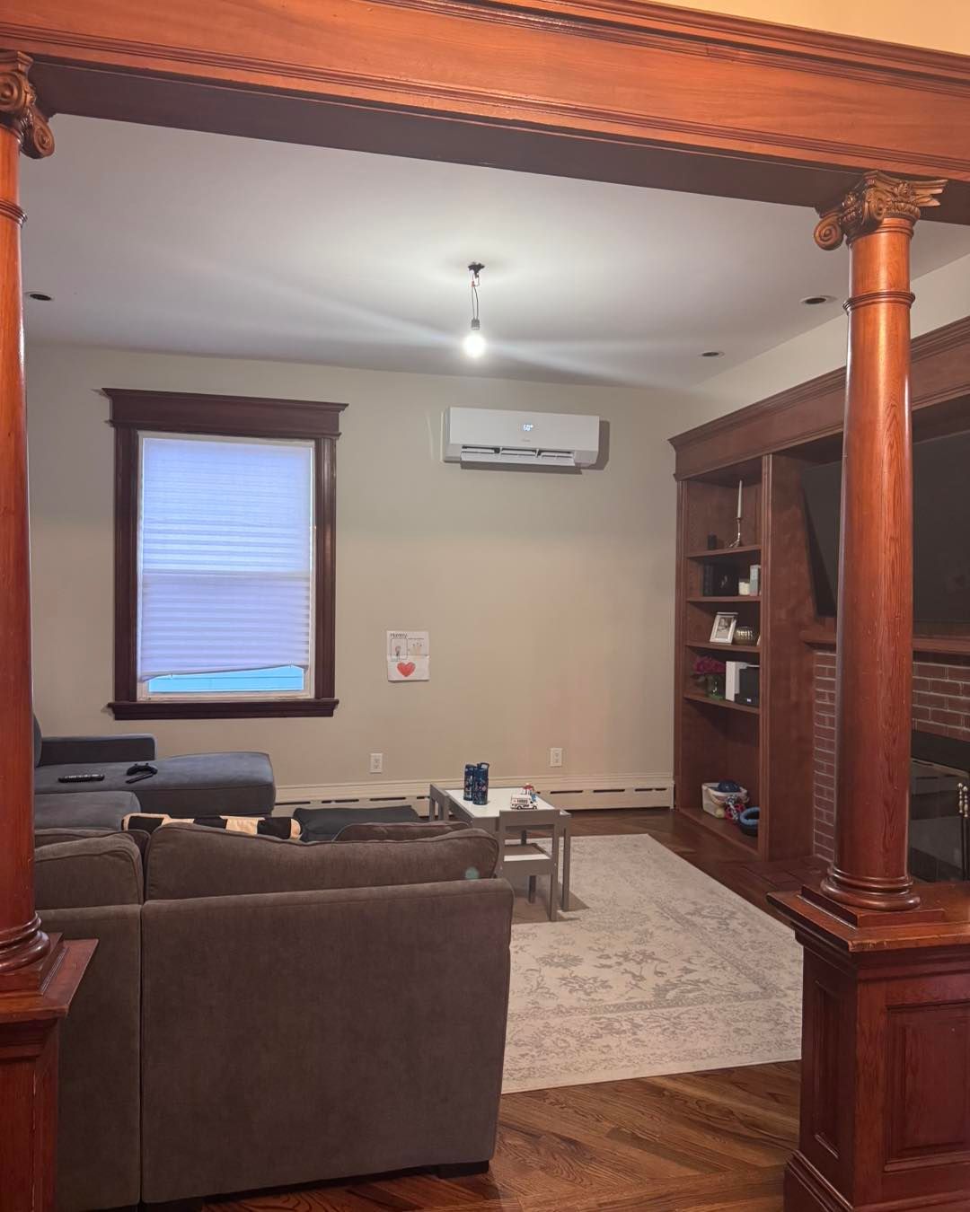 Living room with brown wood columns, built-in shelves, and a gray couch. A window with blinds is on the left.
