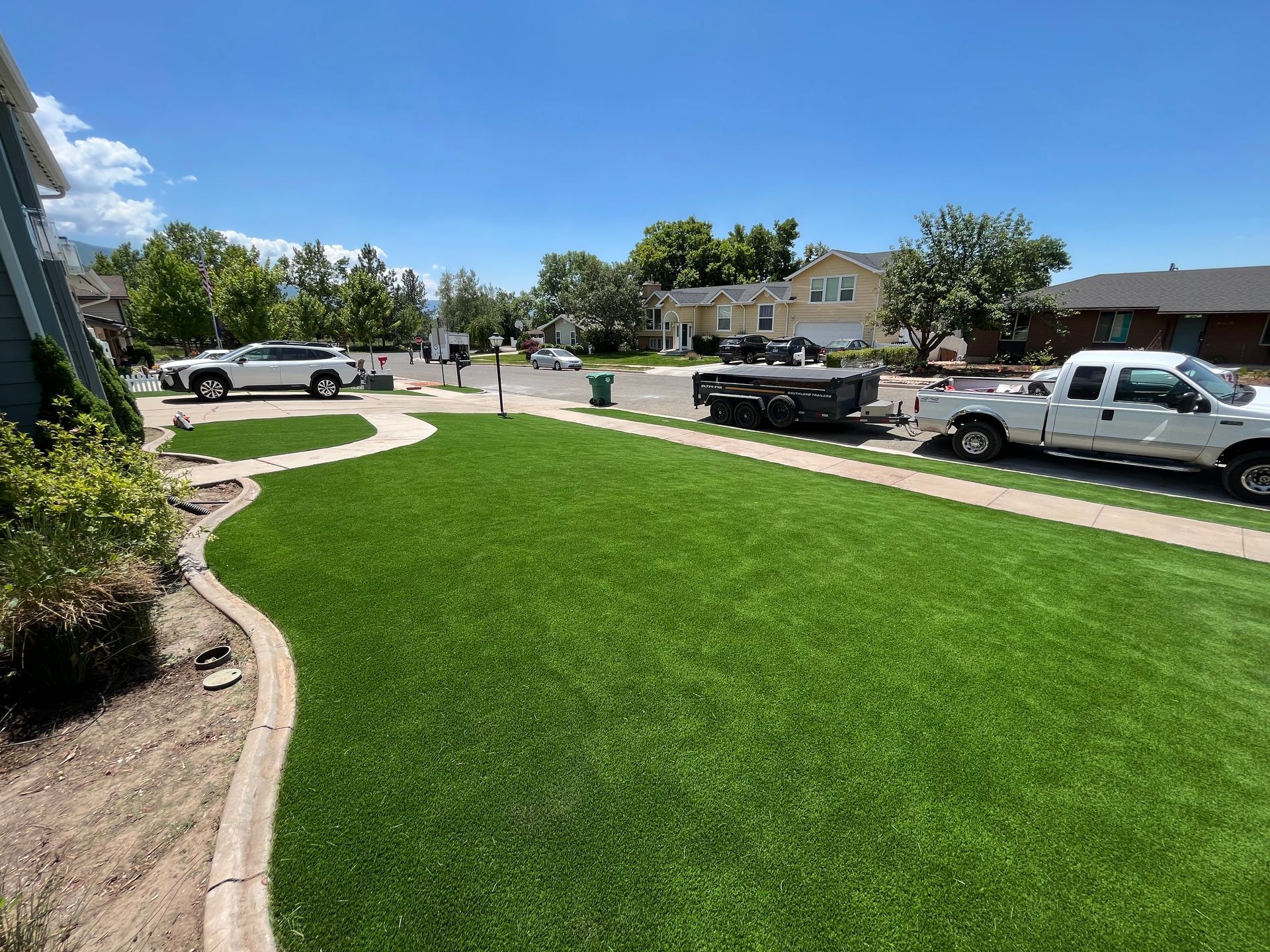 A vibrant green lawn in a suburban neighborhood with white trucks parked nearby on a sunny day.