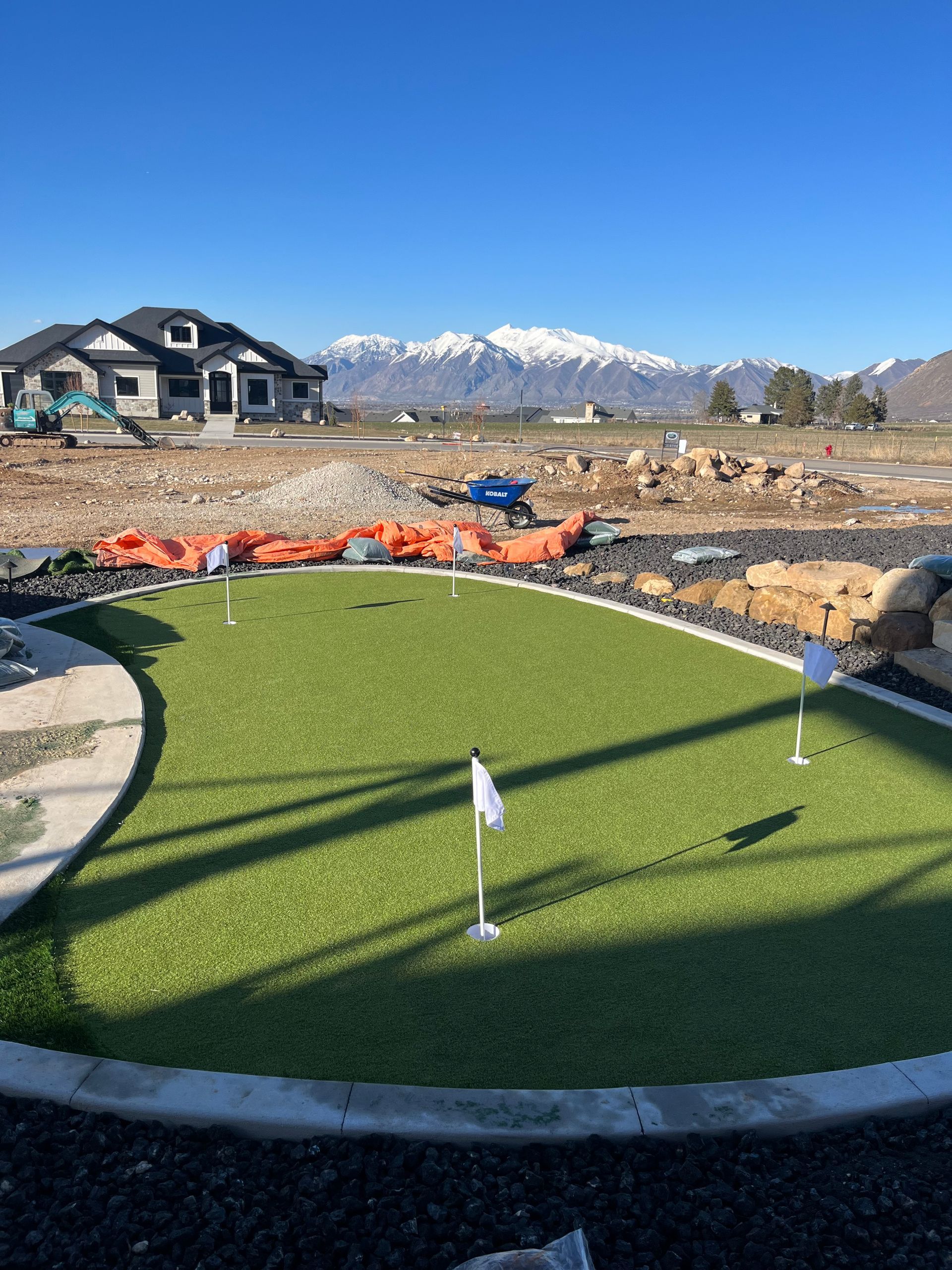 Putting green with white flags, house, mountains, and clear blue sky.