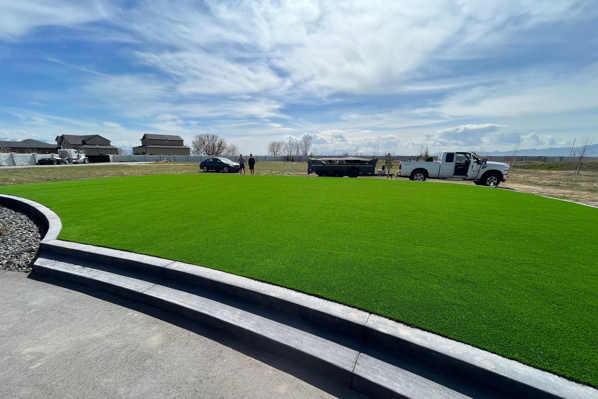 Artificial green lawn with gray curb and white truck under cloudy sky.