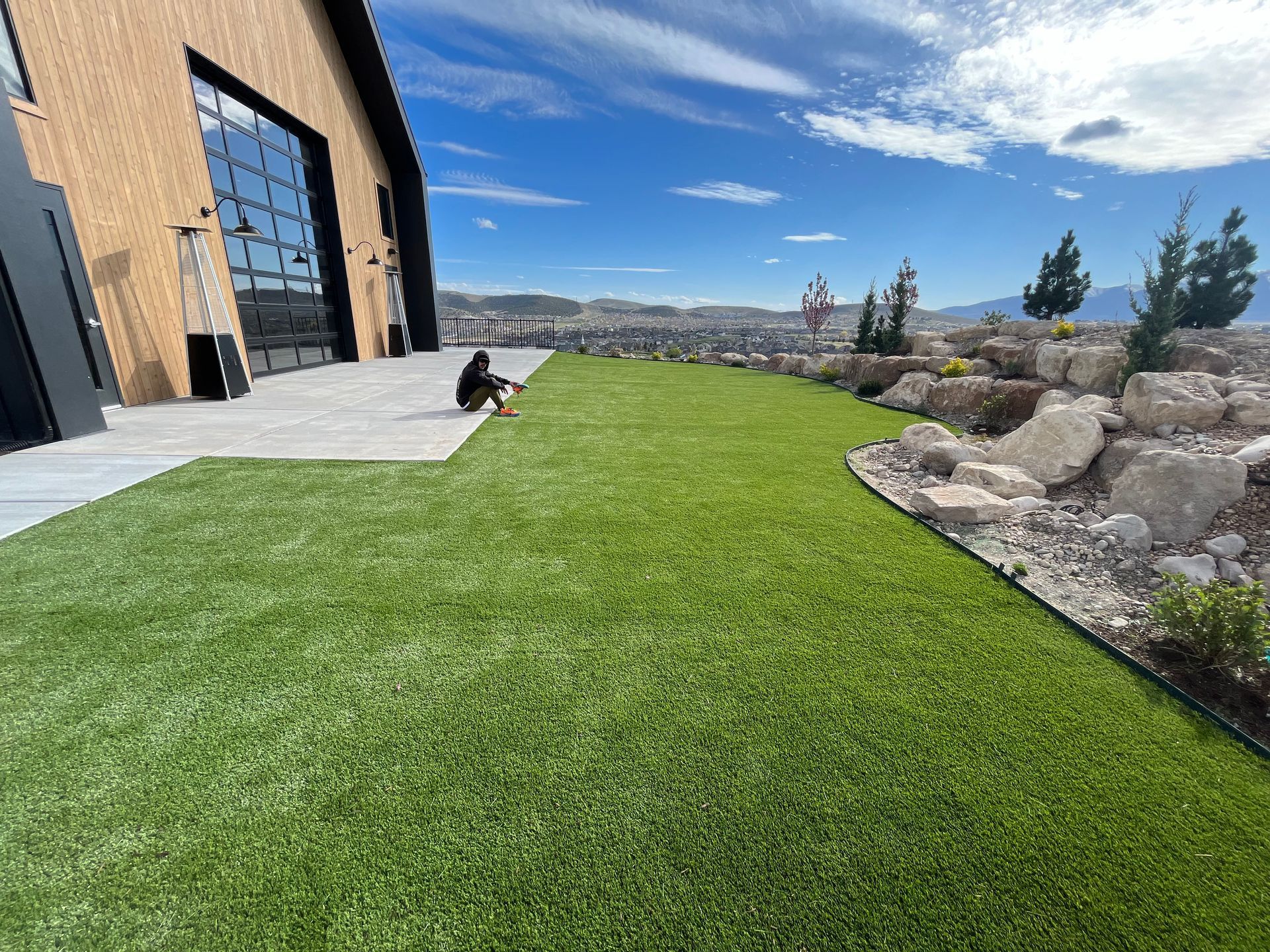 A person sits on a green lawn next to a building. The sky is blue with white clouds.