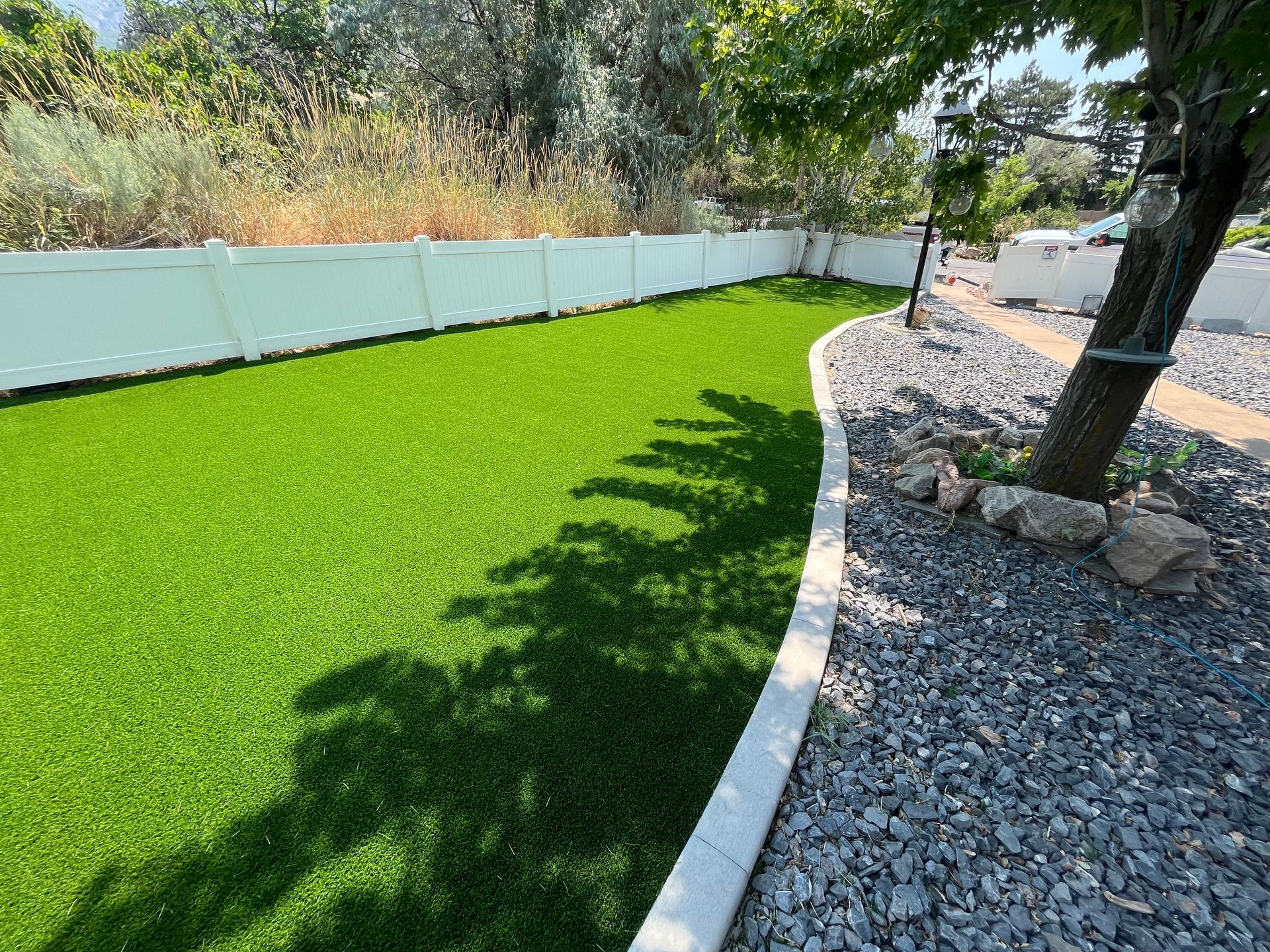 Green artificial turf lawn next to gravel path, bordered by a white fence, under tree shade.