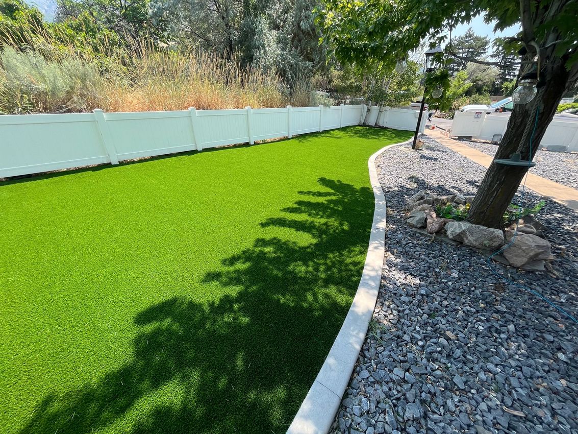 Green artificial turf in a backyard, bordered by a white wall and gravel, shaded by a tree.