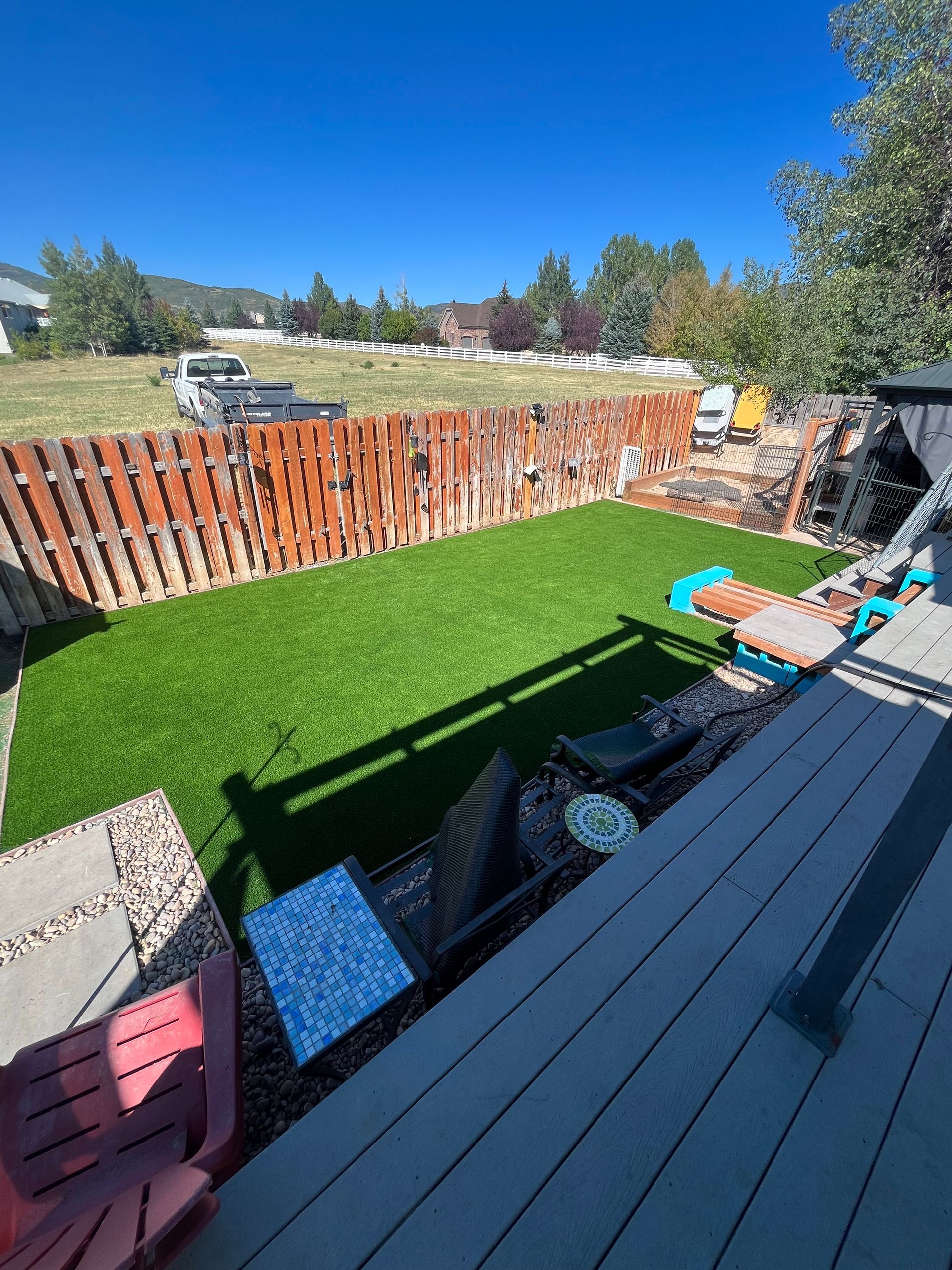 Green turf backyard with a wooden fence, patio furniture, and a sunny blue sky.
