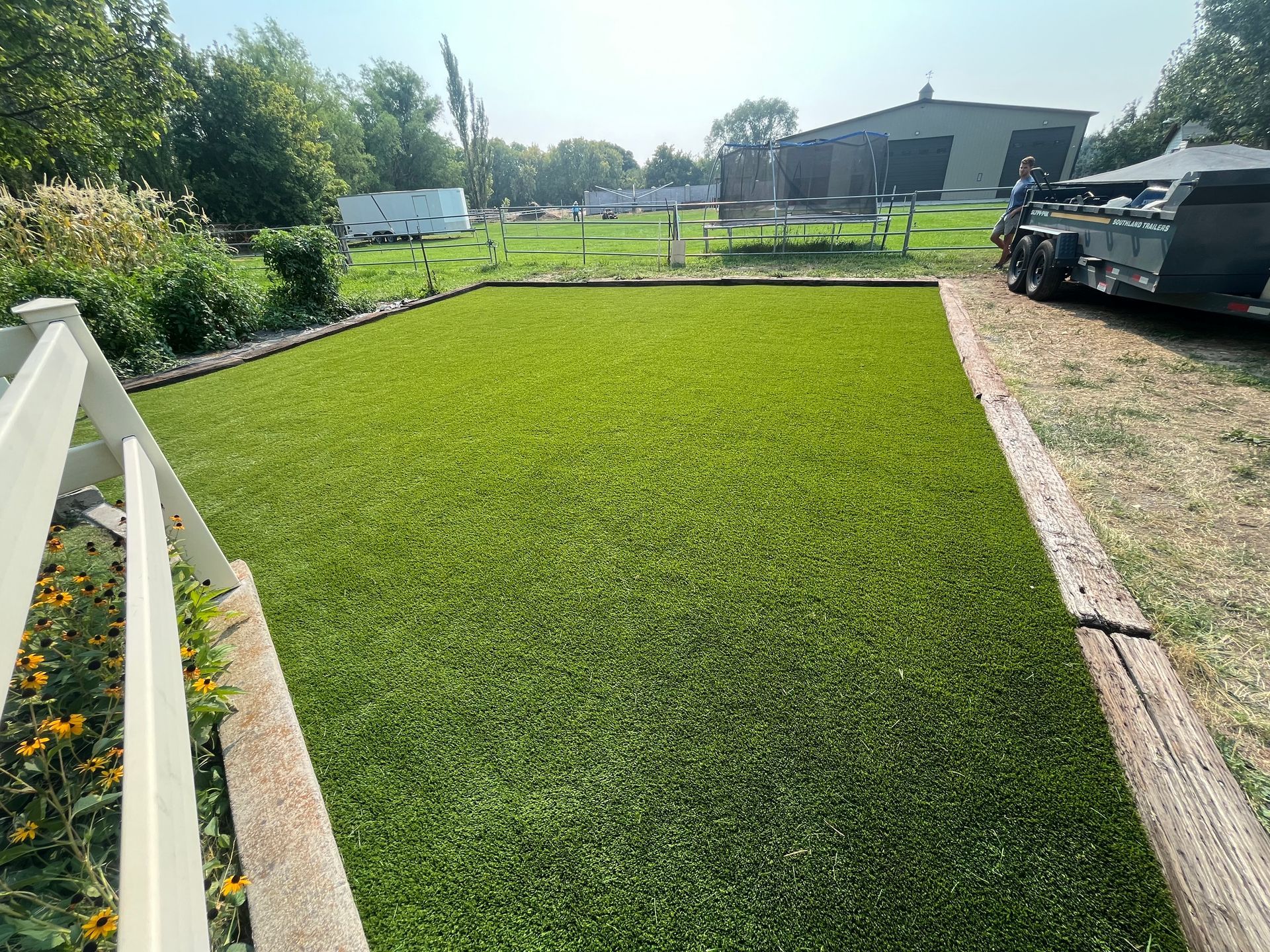 A patch of bright green artificial turf, bordered by wood, with a trailer parked beside it, outdoors.