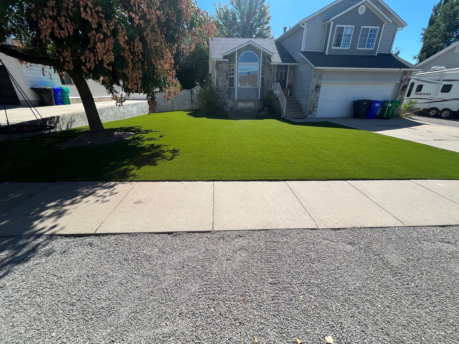 House with green lawn, gray gravel, and sidewalk. Blue bins by the garage, clear sunny day.