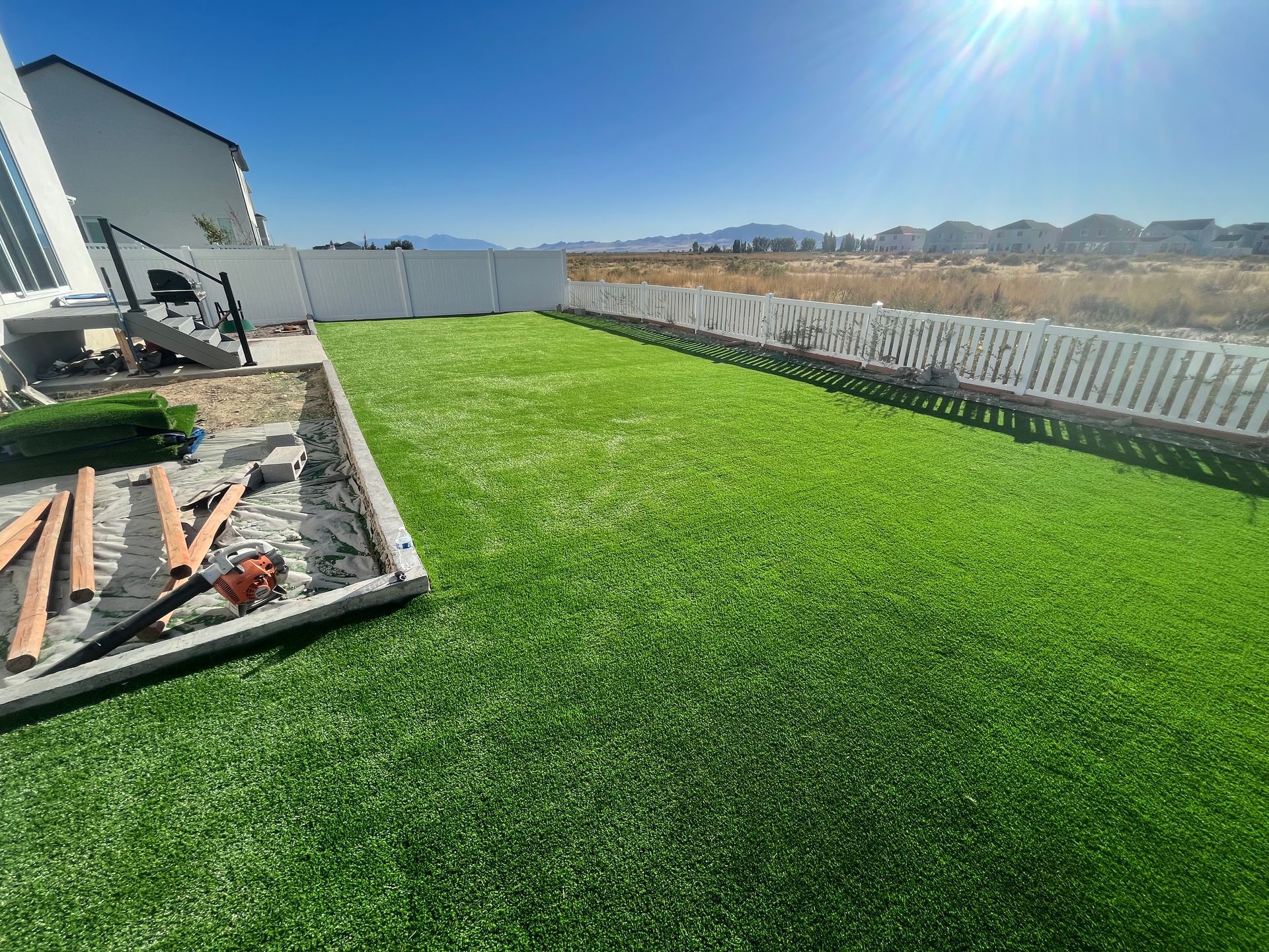 Green artificial turf lawn enclosed by white fence under a bright blue sky.