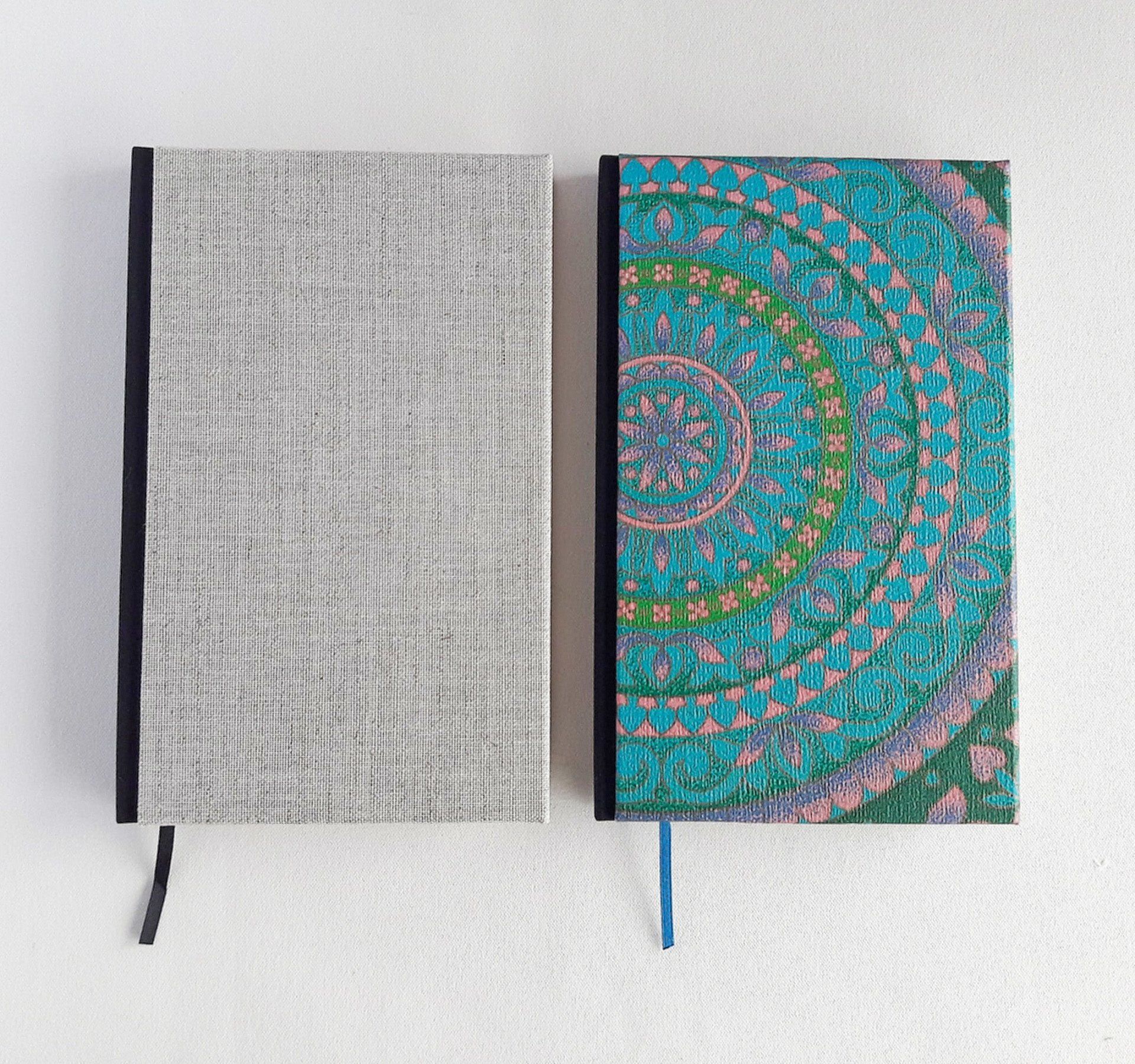 The image shows 2 portrait A5 sized notebooks. The first book on the left is covered in a grey linen fabric with a black spine and black page book mark ribbon. The second book on the right is covered in vintage upcycled wallpaper from the 1960s and 1970s. The patterns are circular and retro in a mandala pattern in light pink purple blue and green. The patterned book has a matching vibrant blue page book mark ribbon. They are laid on a white background.