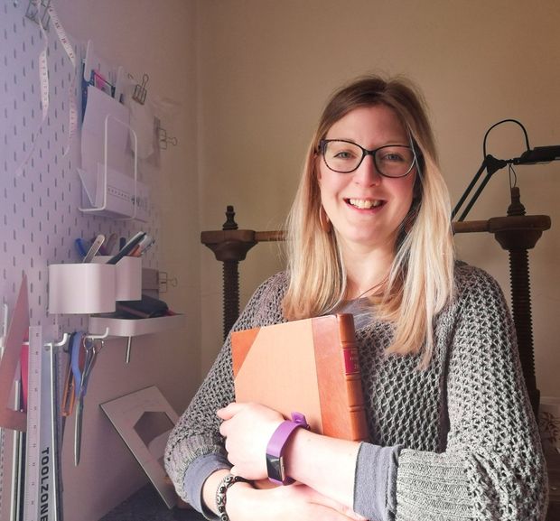 There is a woman called Holly who is the BookBinder of Bespoke Bindings. She is holding a book with a brown leather  spine and cloth covered sides that she has handmade. She stands in front of a large Victorian wooden sewing frame in her studio with her tools hanging on the wall beside her. Holly is smiling and she has long blonde hair and wears glasses. She wears a brown open weave jumper.