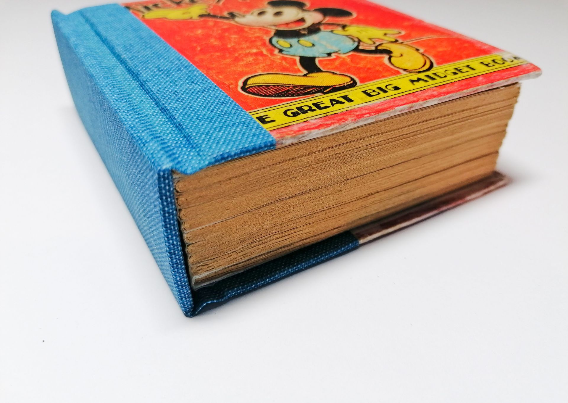 The image shows a small red vintage book from the 1950's with Mickey Mouse on the cover. It is laid on a white background. There is a close up detail from the bottom of the book which shows the spine and neatly trimmed pages within the book, because it has now been repaired and restored. The spine is covered in a blue book cloth and the book has been made into a hardback book.