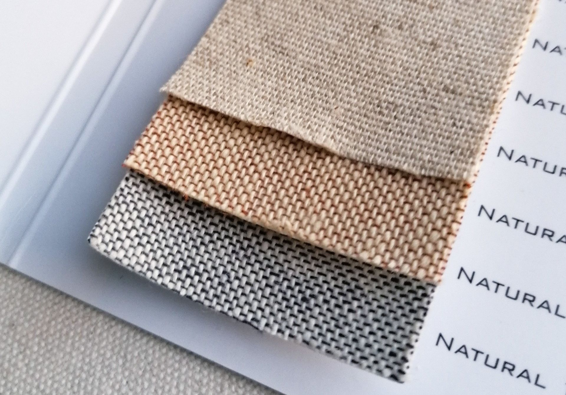 The image shows a collection of 3 natural cottons in a swatch book which can be used as book cloth upon books. The top is a natural light beige colour with flecks of brown. The one underneath is cream with light peach flecks and the one underneath that is cream with dark blue flecks.