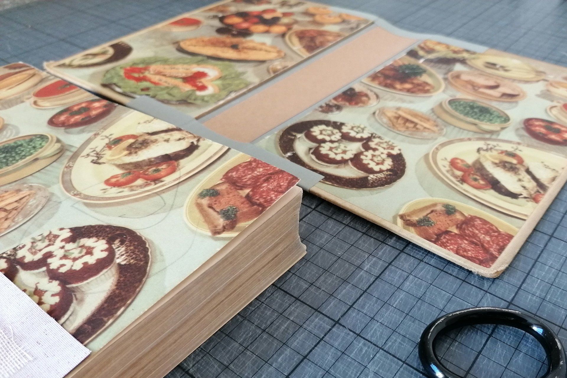An image of a vintage recipe book where it is laid next to the seperate book cover on a workshop cutting mat in the studio of bespoke bindings.