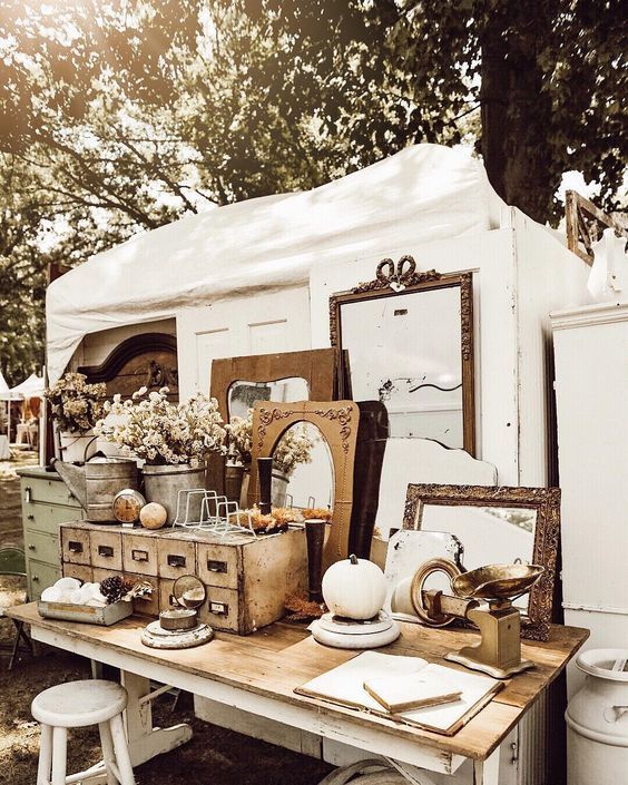 The image shows a vintage stall at a fair. There are many mirrors stacked against one another on the table, most of them have gold frames. There are some old drawers with a vase of pretty flowers on top. There is a white tent behind the stall and a large tree can be seen in the background.
