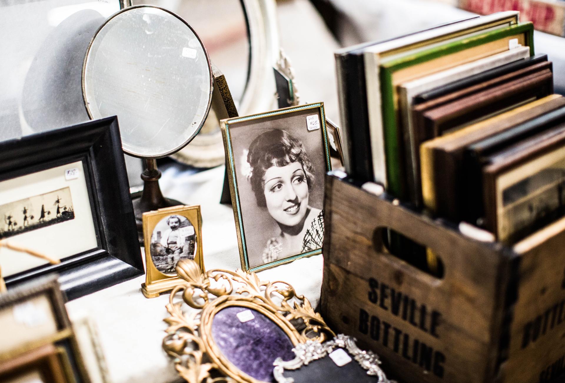 An image of collectables at an antiques fair. They are displayed on a white tablecloth. There is a small frame with a vintage image of a lady from the 1920's, a small ornate gold mirror, an old rustic crate that has Seville Bottling stamped on it. The crate contains more miniature vintage frames.