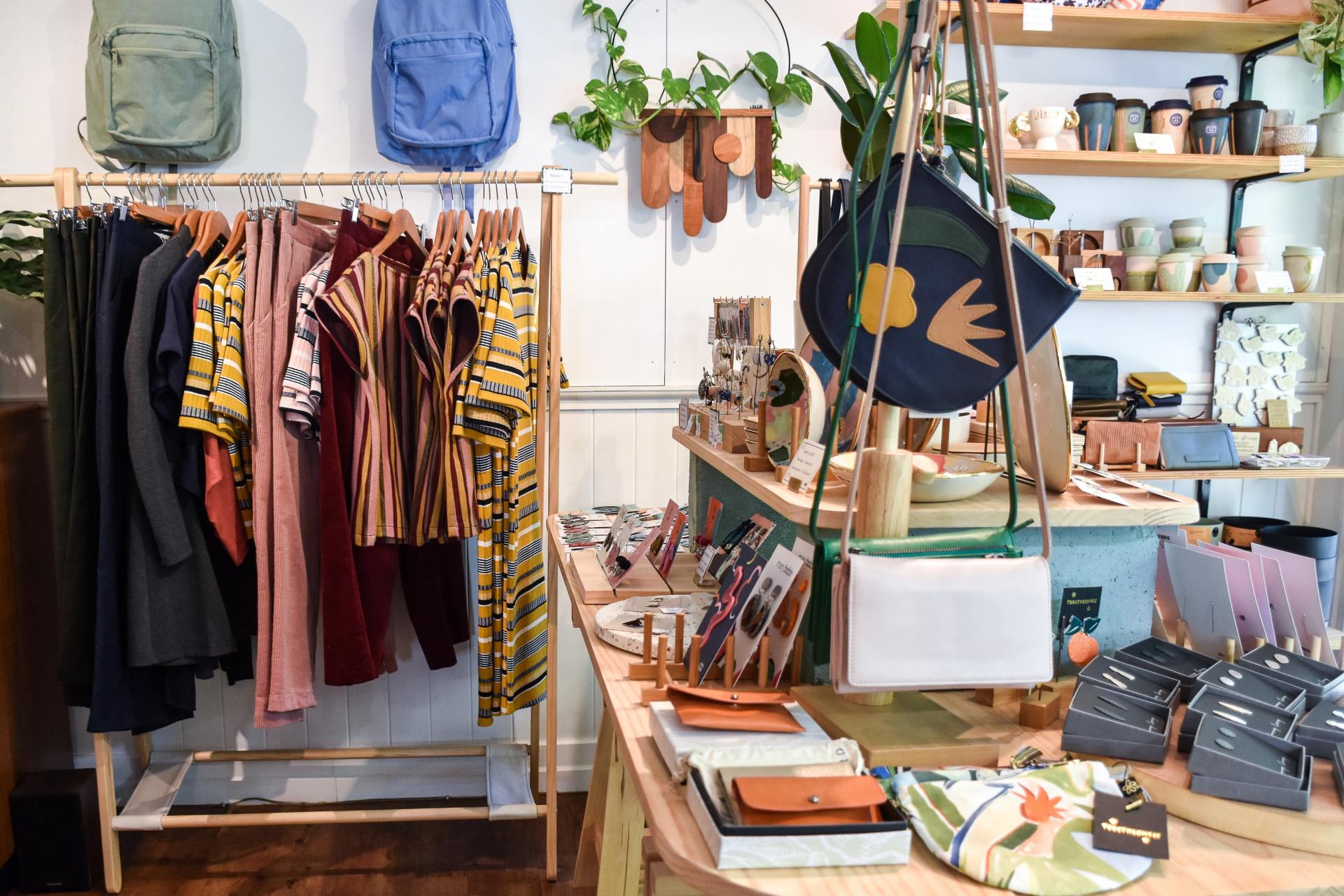 An image of a local boutique gift shop selling handmade goods by local artists and artisans. There is a rail of colourful clothing and 2 rucksacks hanging from the wall one blue and one green. There are some handmade  leather hand bags and purses on display on a table colourful ceramic pots on shelves on the wall in the background.