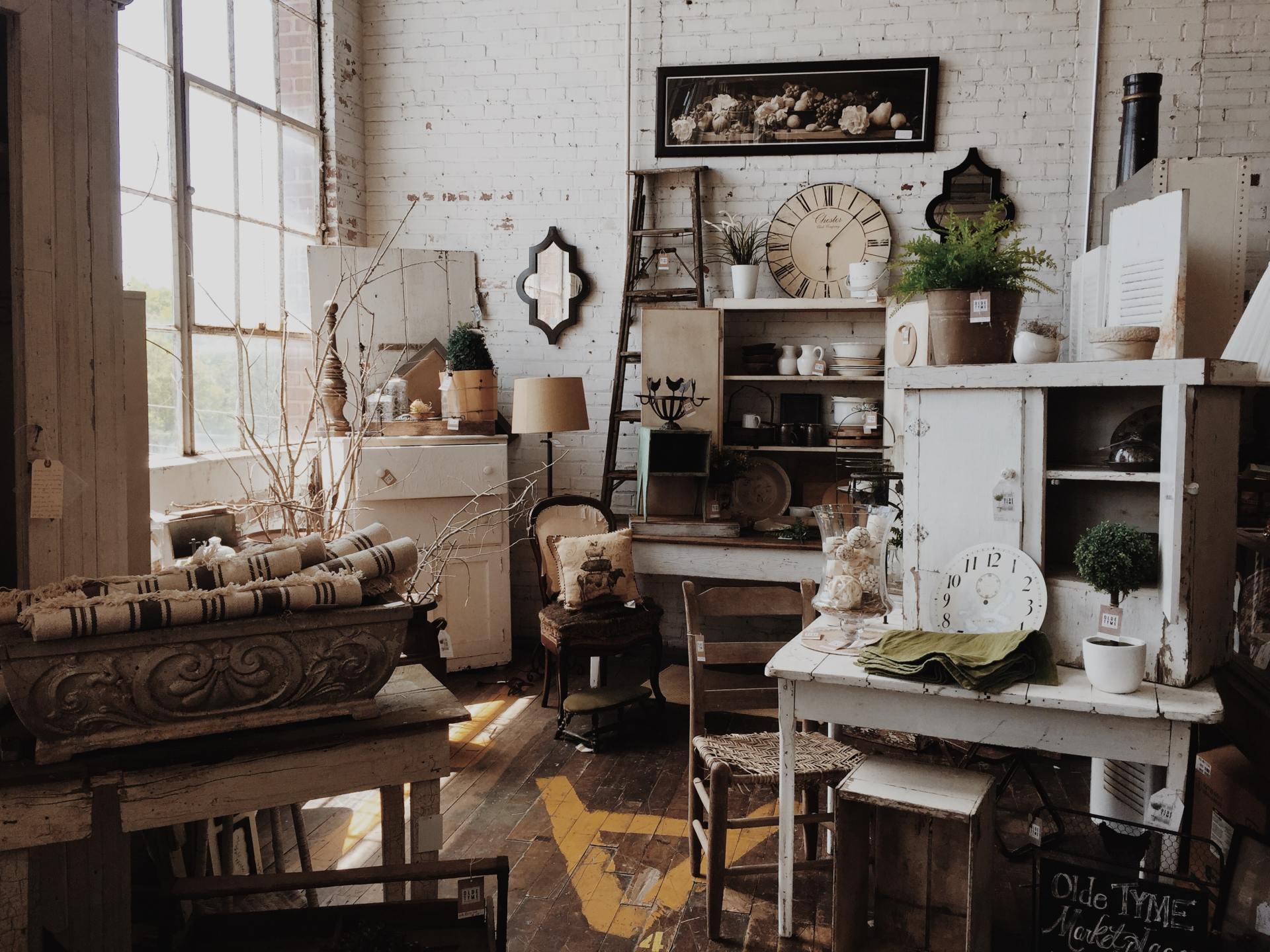 The image shows a vintage homewares store. A large window and white brick wall means the space was one industrial and it may be an old factory. There are various items such as mismatched vintage tables and chairs. The furniture is coloured white, tan brown, and dark brown so they complement one another. There is an old fashioned landscape picture of some flowers which is hanging very high on the white brick wall and an old pair of step ladders and a large cream clock with roman numerals sit next to the picture.