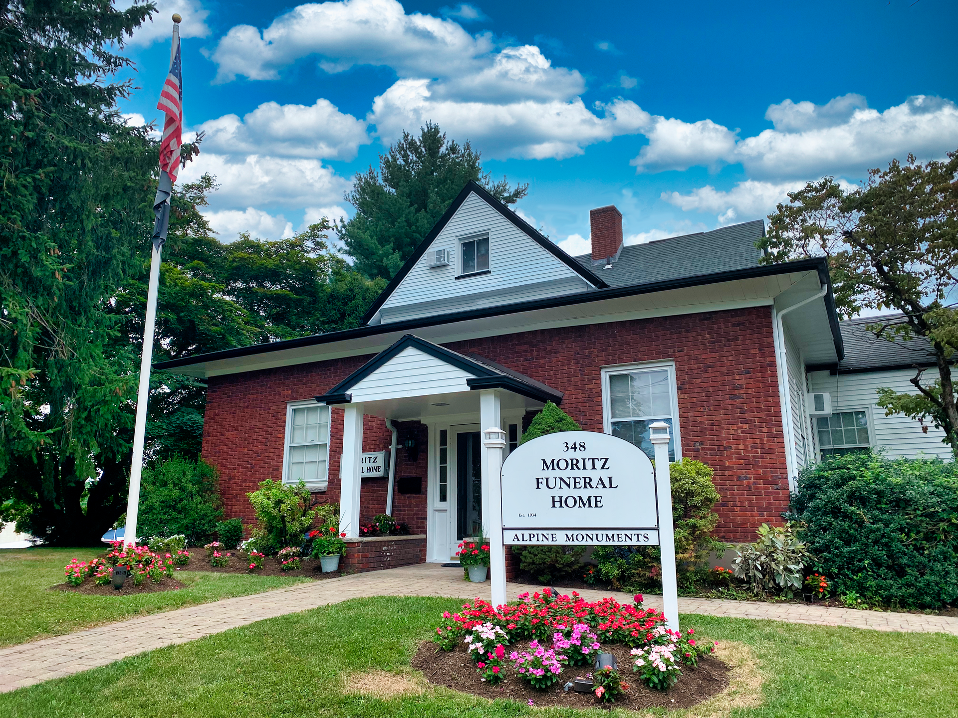 Funeral home with red brick exterior, sign, American flag, and flowers.