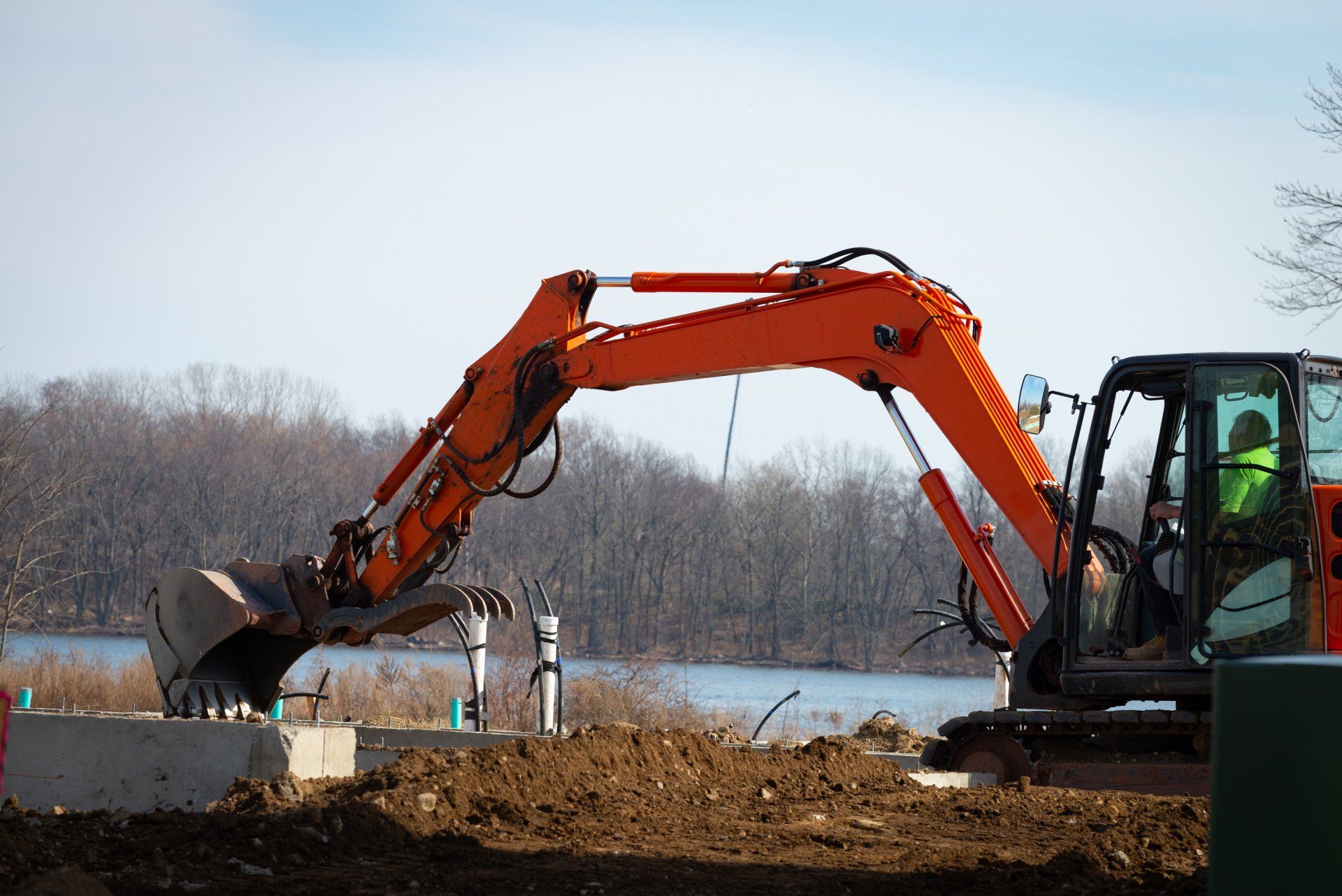 Orange excavator digging earth near a body of water, with a worker visible in the cab.
