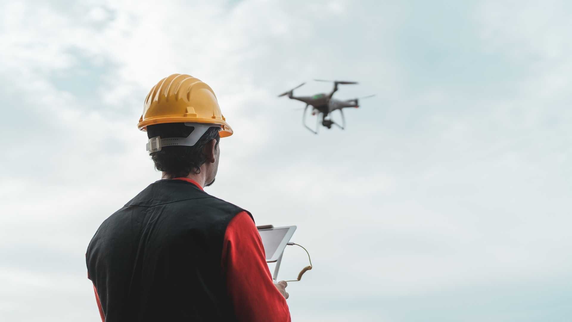 A man in a hard hat is flying a drone while holding a clipboard.