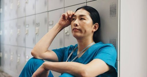A person in blue scrubs sits against lockers, resting a hand on their forehead; weary expression.