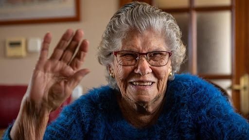 Nurse smiles at computer in hospital room with patient in background.