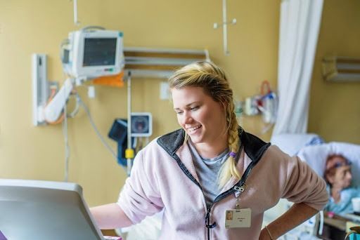 Nurse in a pink jacket smiles while looking at a monitor in a hospital room.