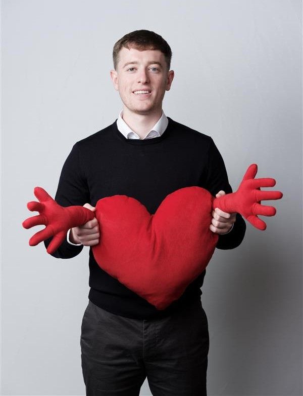 Man in black sweater holding a red heart-shaped pillow with outstretched arms, smiling.
