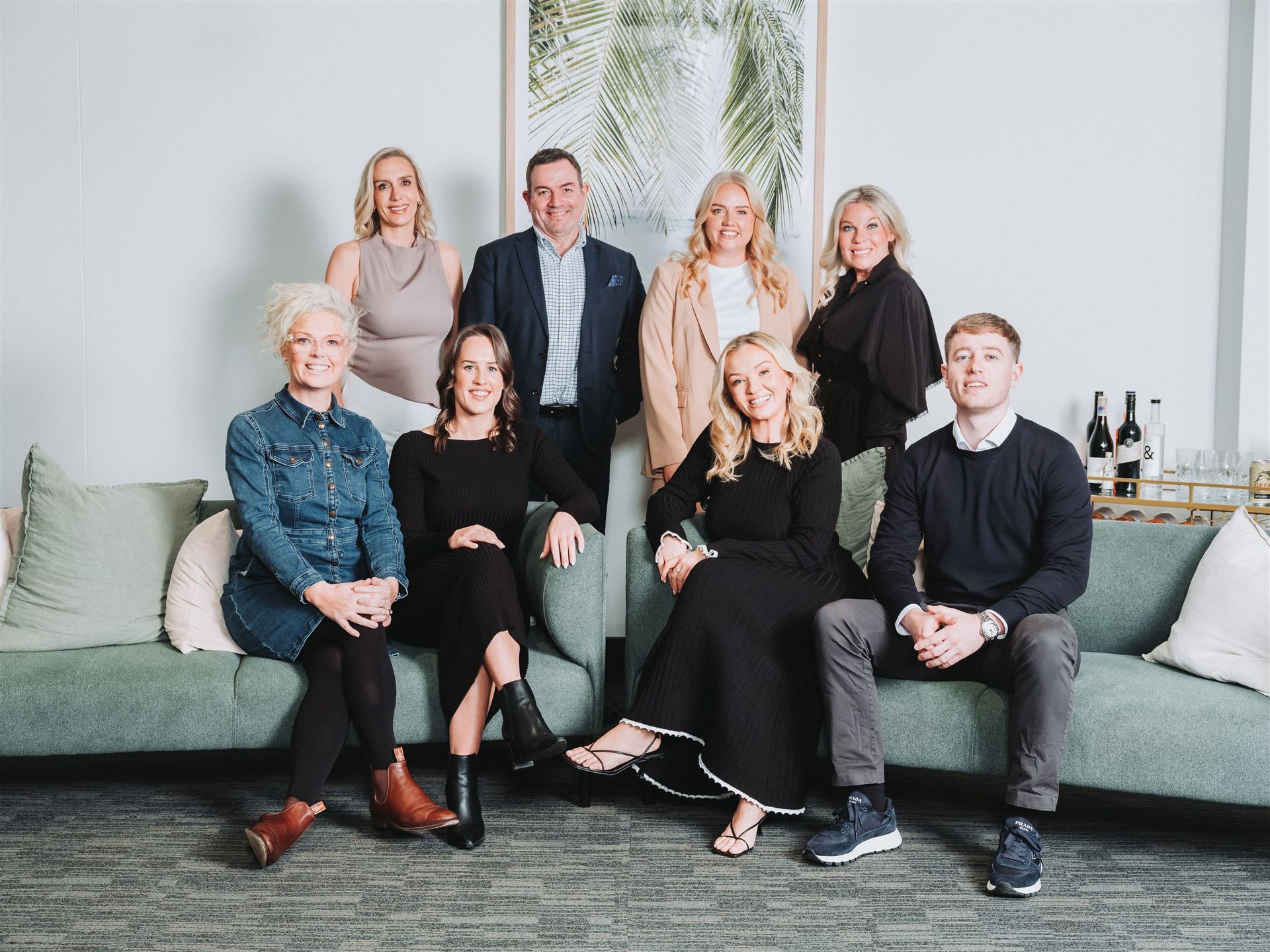 Group of nine people posing on and around a green couch against a white wall; some are smiling.