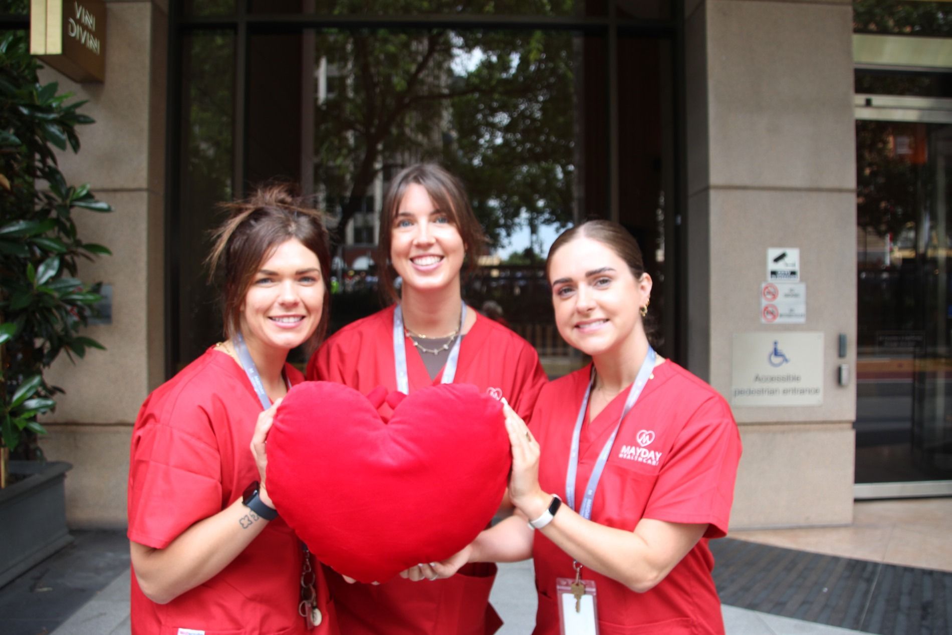 Three people in red scrubs holding a heart-shaped pillow, smiling outside a building.