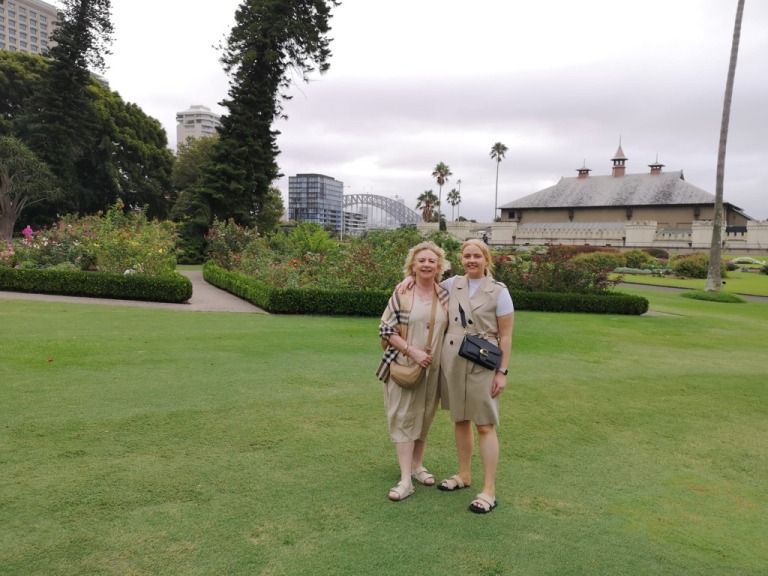 Two women pose in a garden with Sydney Harbour Bridge in the background. Green grass, cloudy sky, and a stone building.