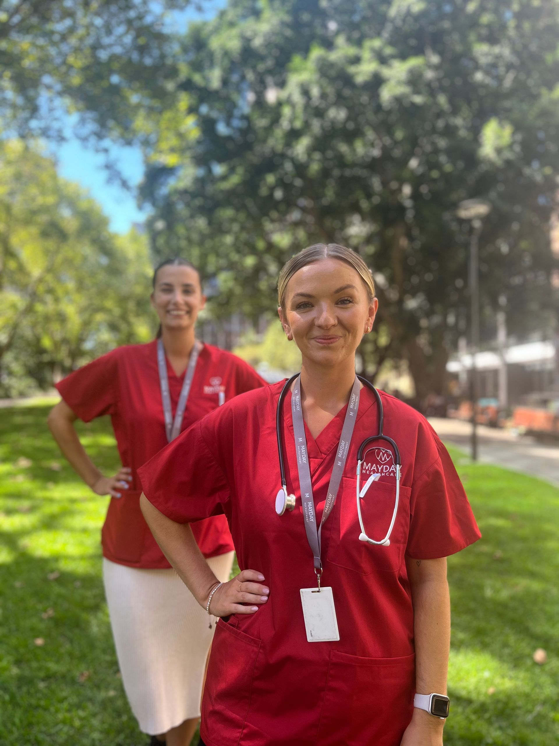 Two healthcare workers in red scrubs, outdoors with stethoscope and hands on hips.