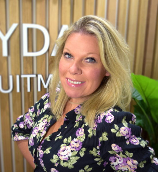 Blonde woman smiling, wearing a floral top, in front of a wooden wall with the word 