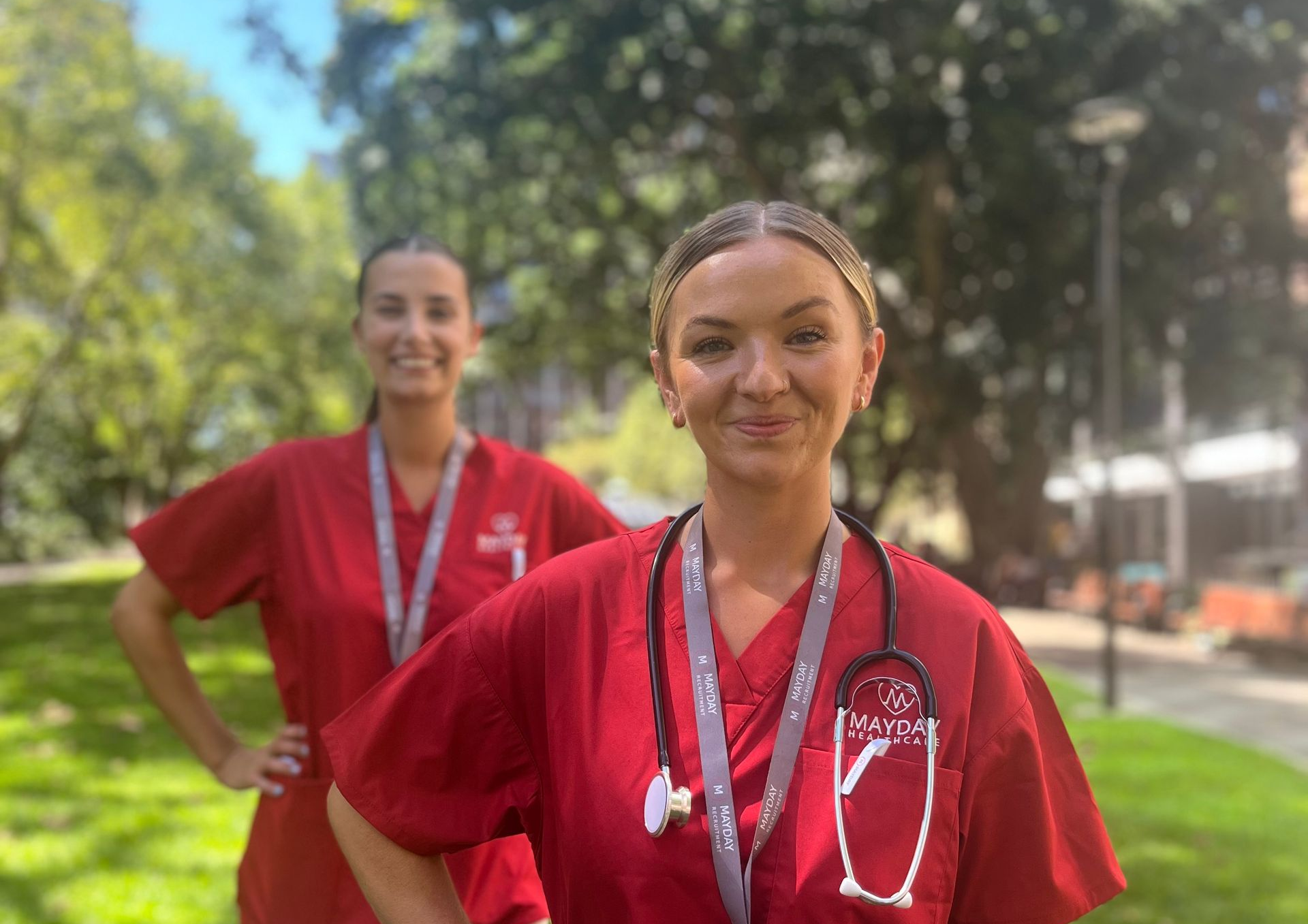 Two healthcare workers in red scrubs, outdoors with stethoscope and hands on hips.