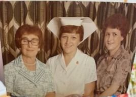 Three women posing for a photo. A woman in a nurse's uniform is centered. The other two women are in casual wear. Floral curtain backdrop.