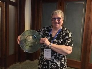 Woman holding an award, smiling. She wears glasses, a floral top, and a lanyard. Interior setting with wooden doors.