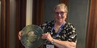 Three women posing for a photo. A woman in a nurse's uniform is centered. The other two women are in casual wear. Floral curtain backdrop.