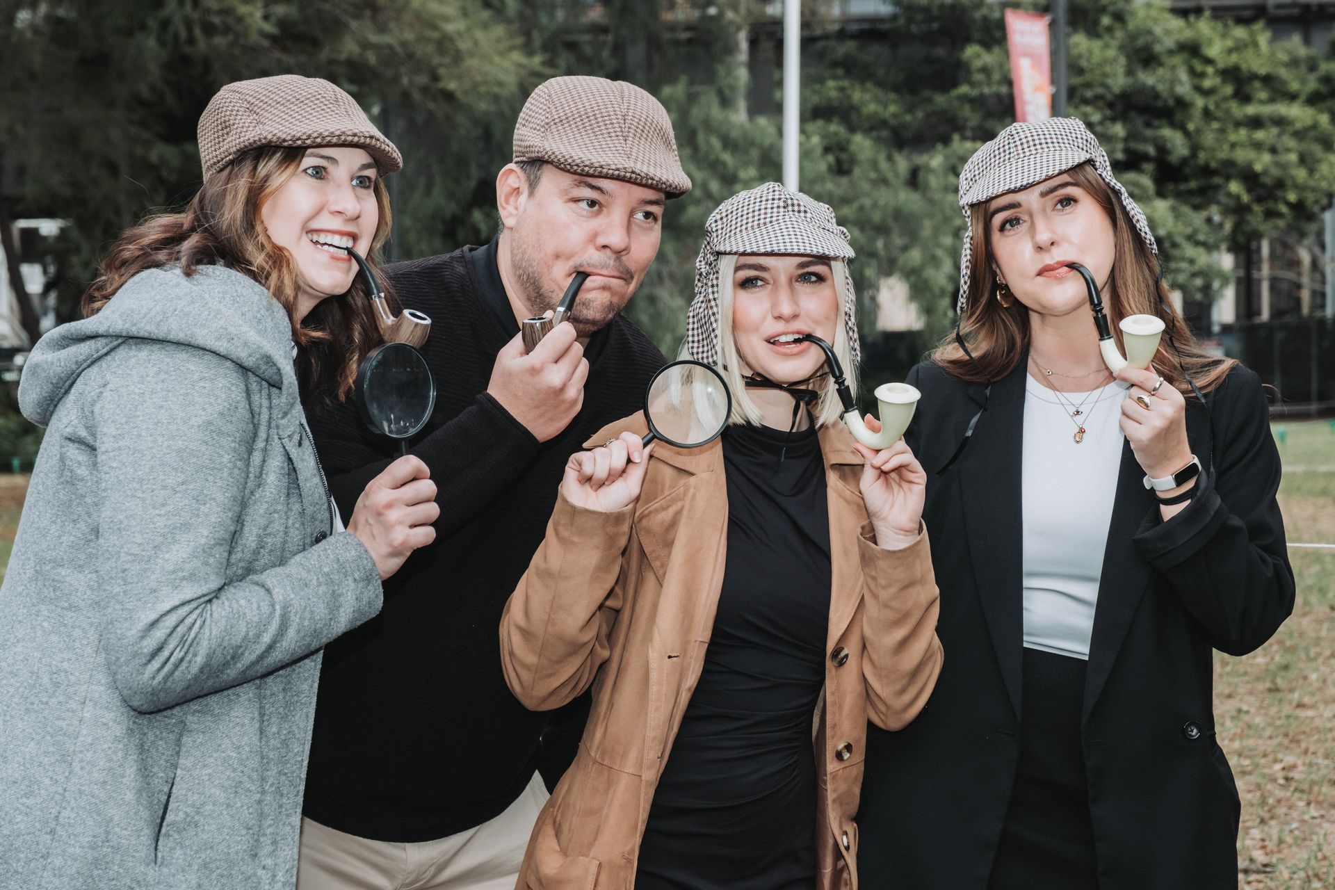Four people in detective costumes: holding pipes and magnifying glasses. Outdoors.