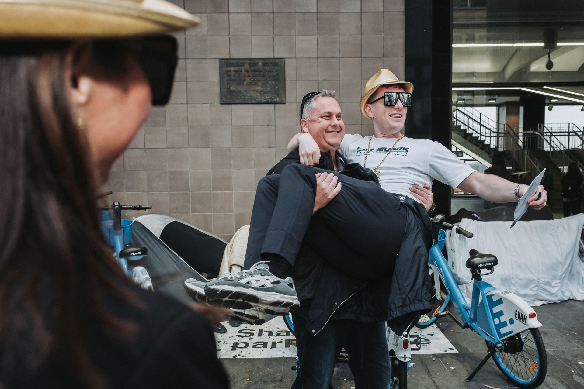 Man holding another man up, taking a selfie. Person watches. Blue bikes and building background.